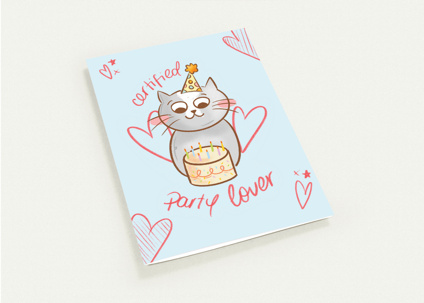 A square greeting card featuring a cute cartoon cat wearing a party hat and holding a birthday cake, with the words 'cutefied party lover' and heart illustrations on a pale blue background, photographed on a light neutral surface.