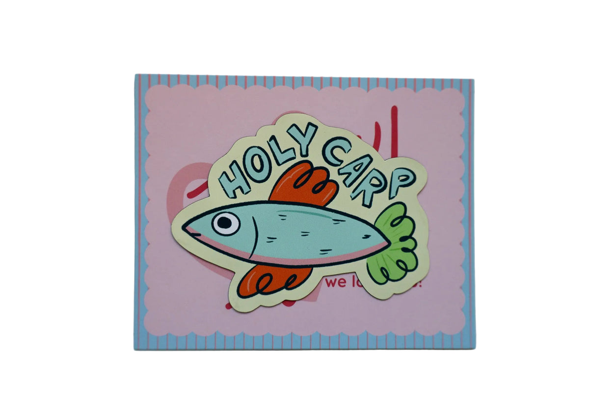A handmade paper craft sticker or card featuring a cartoon fish with the text 'HOLY CARP' on a pink background with a blue scalloped border.
