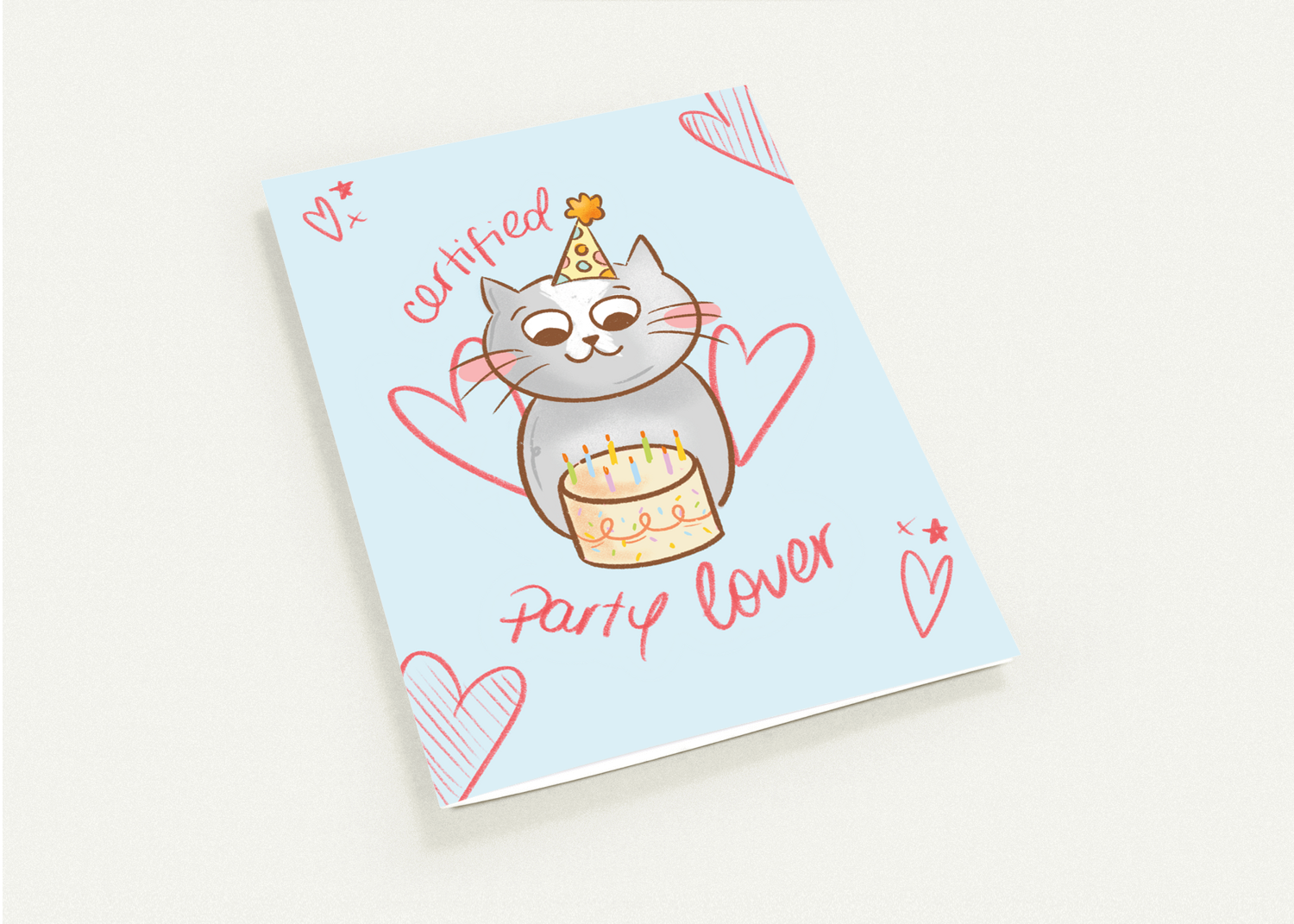 A square greeting card featuring a cute cartoon cat wearing a party hat and holding a birthday cake, with the words 'cutefied party lover' and heart illustrations on a pale blue background, photographed on a light neutral surface.
